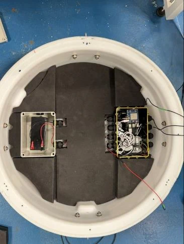 Top-down view inside the buoy shell showing the two IP68 enclosures, battery on the left and electronics and communications on the right, mounted on the closed-cell foam deck.